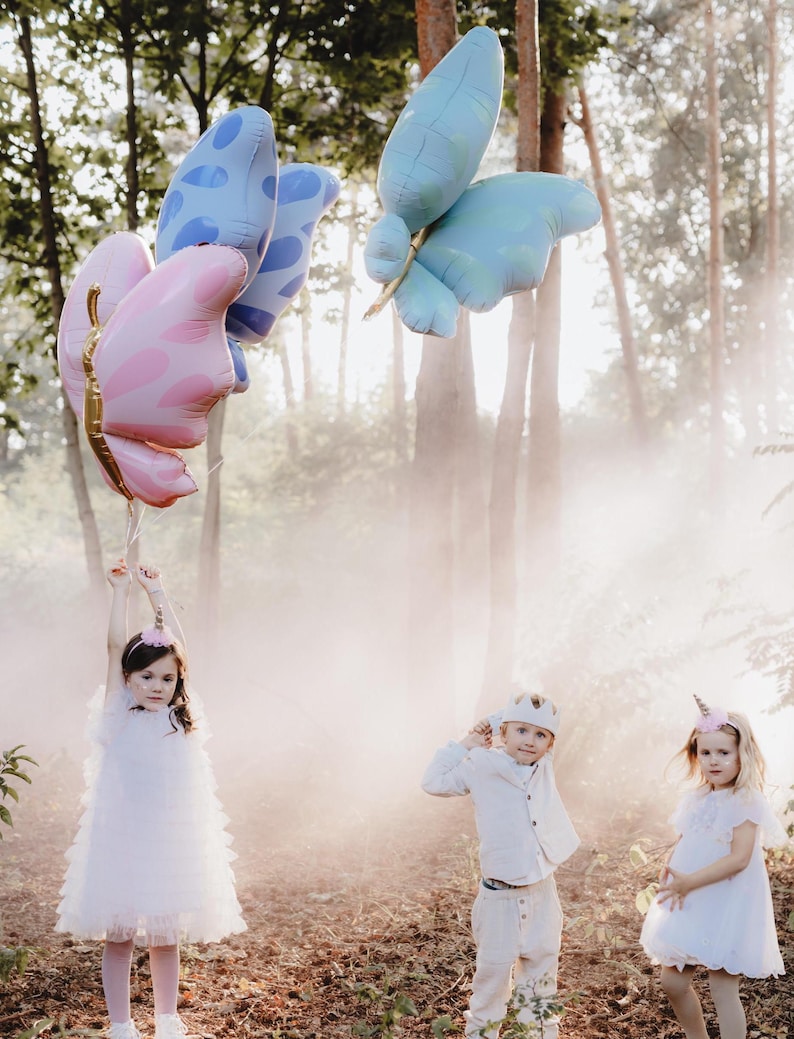 May include: Three children dressed in white clothing and wearing unicorn and crown headbands play in a foggy forest with three large butterfly balloons. The balloons are pink, blue, and blue with white polka dots.