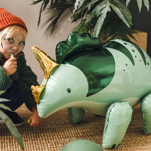 Puede incluir: Un ni&ntilde;o con un sombrero rojo y una chaqueta verde mira a un gran dinosaurio inflable verde con una lupa. El dinosaurio tiene cuernos dorados y un cuerpo con estampado verde y blanco.
