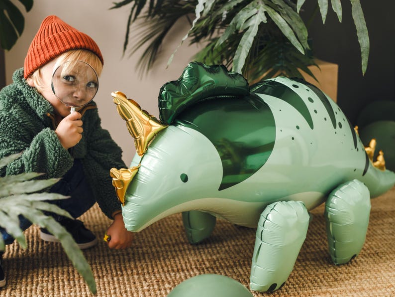Puede incluir: Un ni&ntilde;o con un sombrero rojo y una chaqueta verde mira a un gran dinosaurio inflable verde con una lupa. El dinosaurio tiene cuernos dorados y un cuerpo con estampado verde y blanco.