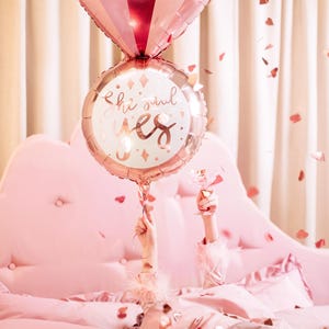 May include: A pink and gold foil balloon shaped like a ring with the words "she said yes" on it. The balloon is held up by a person's hand in a pink bed with confetti.