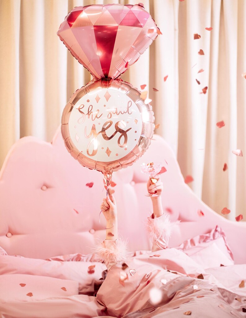 May include: A pink and gold foil balloon shaped like a ring with the words "she said yes" on it. The balloon is held up by a person's hand in a pink bed with confetti.
