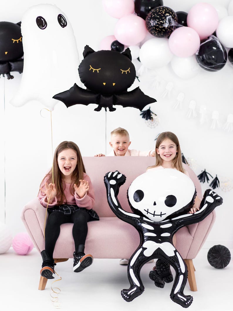 Kids Halloween party with skeleton, ghost, and bat foil balloons by PartyDeco; playful decor with black, white, and pink balloon garland and smiling children on pink sofa