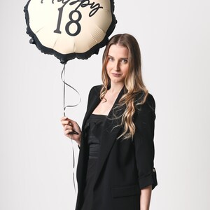 May include: A woman in a black blazer holds a gold and black balloon with the text "Happy 18" printed on it.