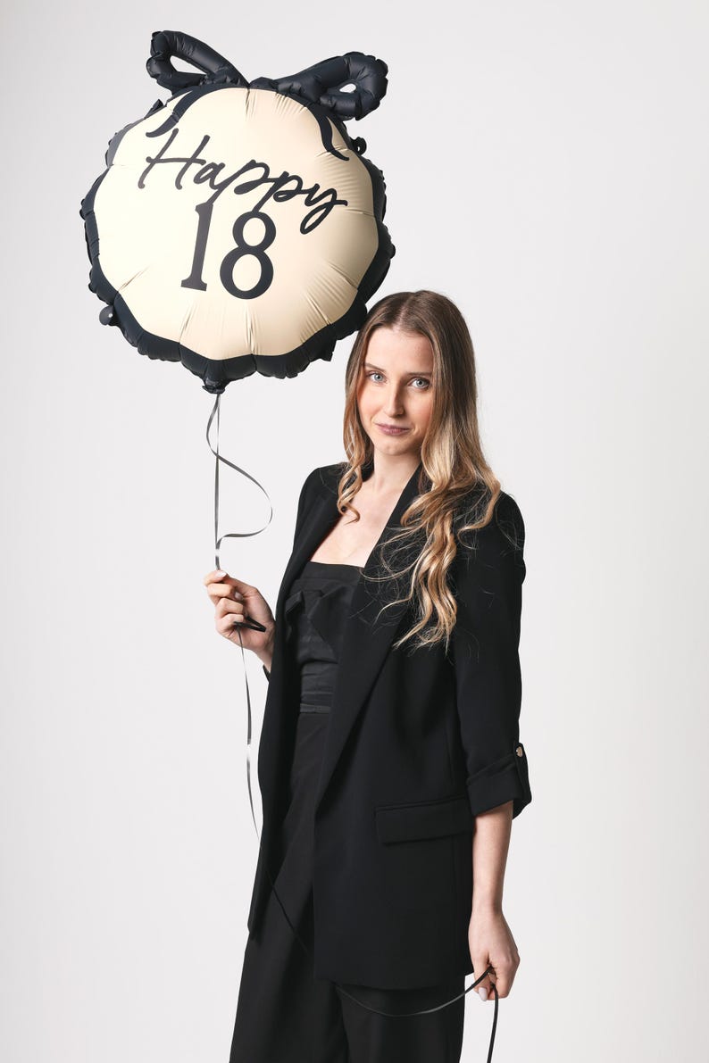 May include: A woman in a black blazer holds a gold and black balloon with the text "Happy 18" printed on it.