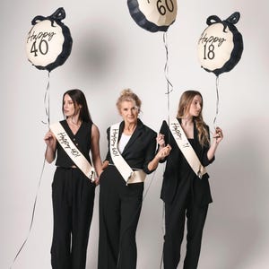 May include: Three women wearing black outfits and sashes with the text "Happy 40!"  "Happy 60!" and "Happy 18!"  They are holding black and white balloons with the text "Happy 40", "Happy 60", and "Happy 18".