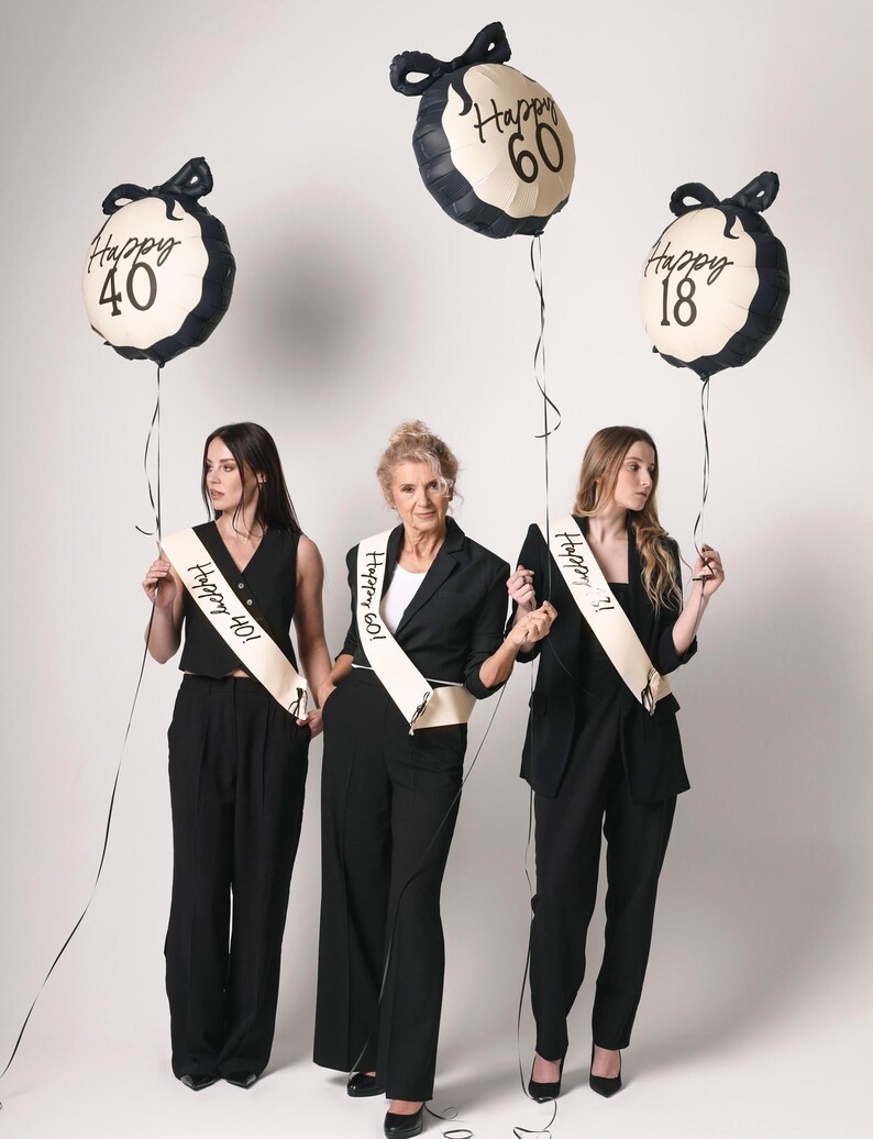 May include: Three women wearing black outfits and sashes with the text "Happy 40!"  "Happy 60!" and "Happy 18!"  They are holding black and white balloons with the text "Happy 40", "Happy 60", and "Happy 18".
