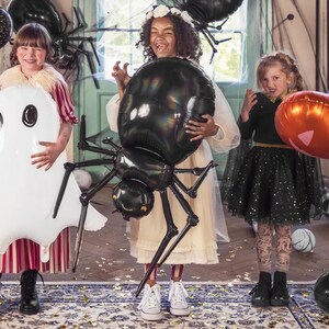 Op de afbeelding: Drie kinderen verkleed voor Halloween poseren met zwarte, witte en oranje ballonnen. Het meisje links houdt een witte spookballon vast. Het meisje in het midden houdt een zwarte spinballon vast. Het meisje rechts houdt een oranje pompoenballon vast.