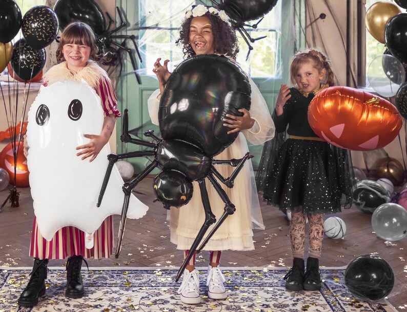 Op de afbeelding: Drie kinderen verkleed voor Halloween poseren met zwarte, witte en oranje ballonnen. Het meisje links houdt een witte spookballon vast. Het meisje in het midden houdt een zwarte spinballon vast. Het meisje rechts houdt een oranje pompoenballon vast.