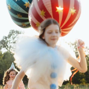 May include: Two large, colorful balloons with star patterns float above two children running through a grassy field. One balloon is red and white striped, the other is blue with stripes. The children are wearing festive outfits.