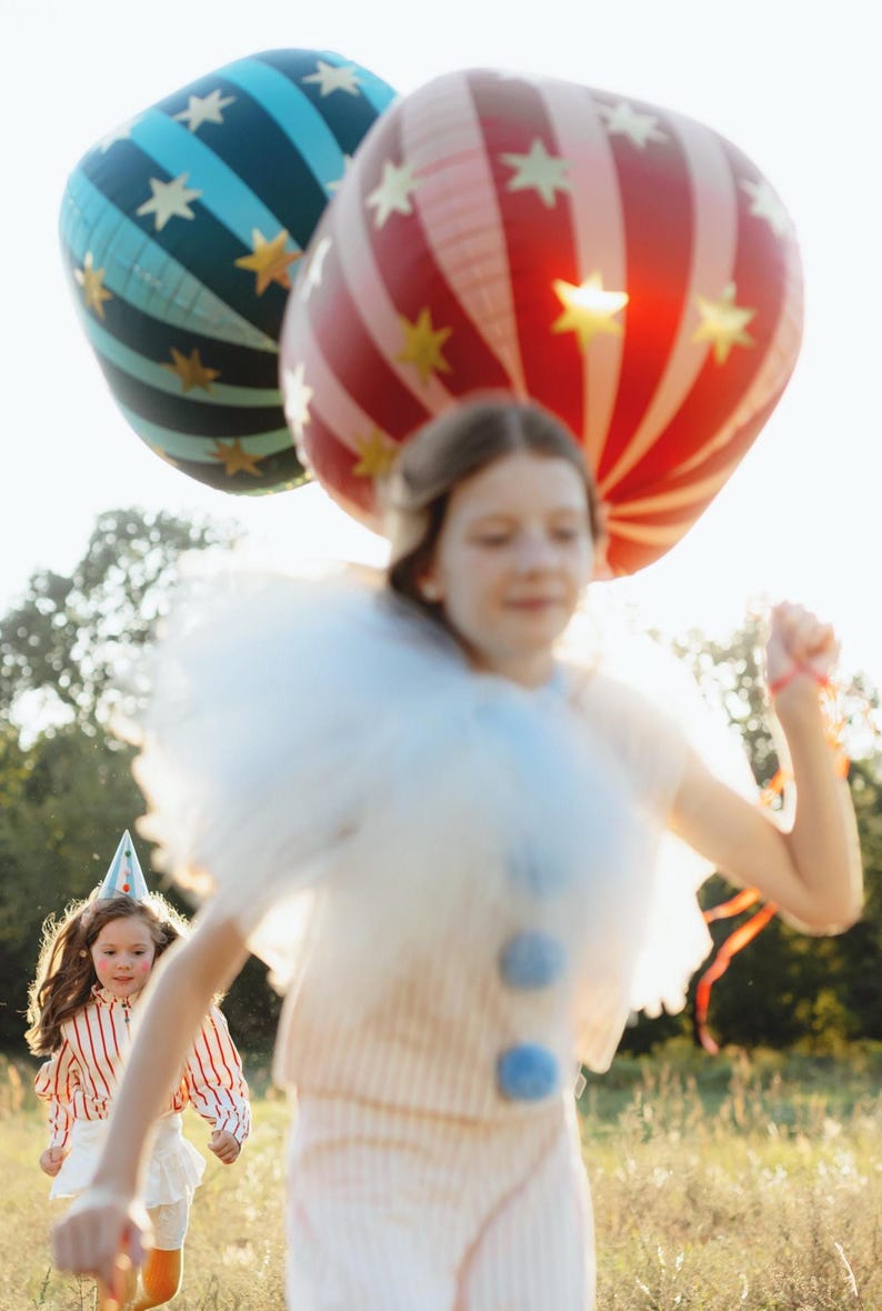 May include: Two large, colorful balloons with star patterns float above two children running through a grassy field. One balloon is red and white striped, the other is blue with stripes. The children are wearing festive outfits.