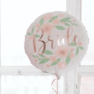 May include: A round white balloon with the word "Bride" in rose gold script. The balloon is decorated with a floral wreath of pink flowers and green leaves, perfect for a bridal shower or wedding celebration.