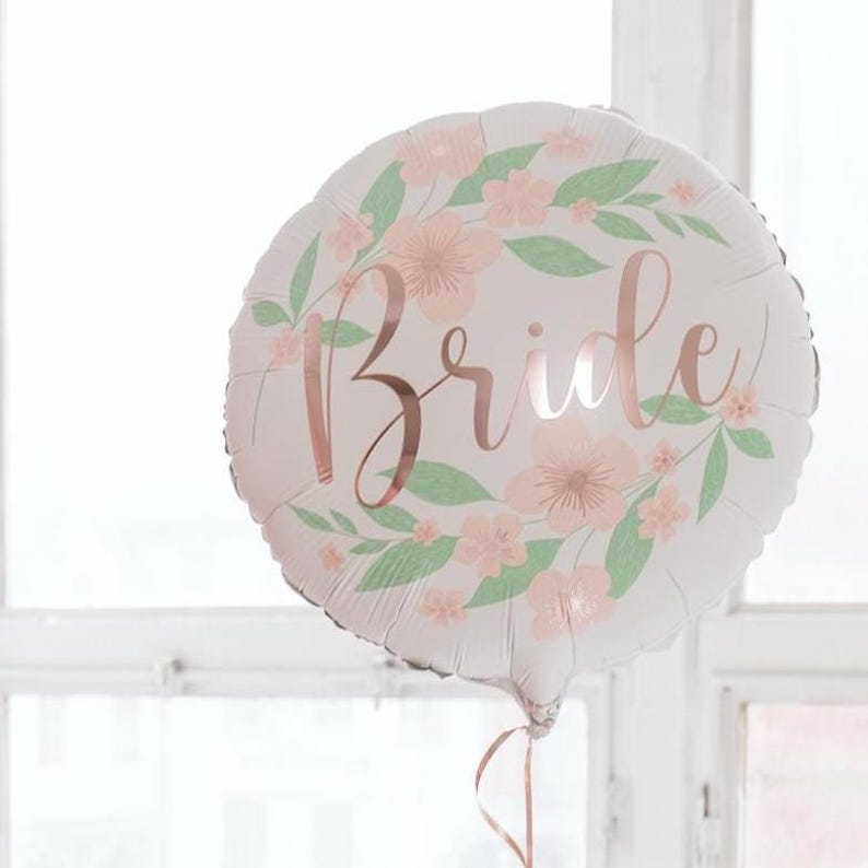 May include: A round white balloon with the word "Bride" in rose gold script. The balloon is decorated with a floral wreath of pink flowers and green leaves, perfect for a bridal shower or wedding celebration.