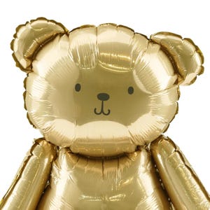 May include: A large, gold-colored teddy bear balloon. The balloon has a simple, cartoon-like face with black eyes and a small, black nose. The bear's ears are rounded, and the balloon is inflated, with visible seams.