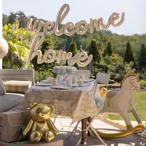 Op de afbeelding: Een babyshower-sc&egrave;ne met een gouden teddybeerballon, een schommelpaardballon en een tafel gedekt met bloemig servies. Een ballonnenbanner spelt "welcome home", en een ronde ballon zegt "Oh Baby".