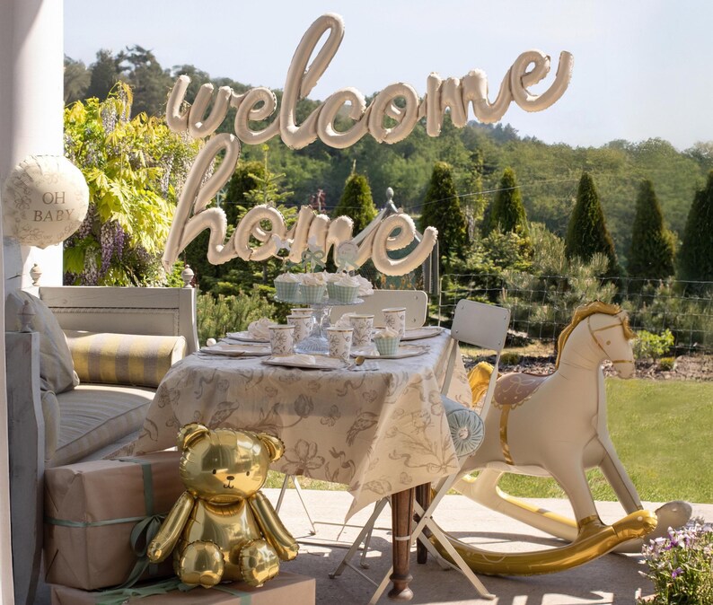 Op de afbeelding: Een babyshower-sc&egrave;ne met een gouden teddybeerballon, een schommelpaardballon en een tafel gedekt met bloemig servies. Een ballonnenbanner spelt "welcome home", en een ronde ballon zegt "Oh Baby".