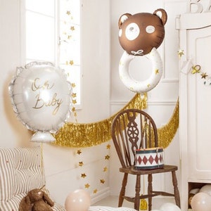 May include: A white balloon with the text "Oh Baby" in gold lettering, a brown bear balloon with a white ring around it, and gold fringe garland.
