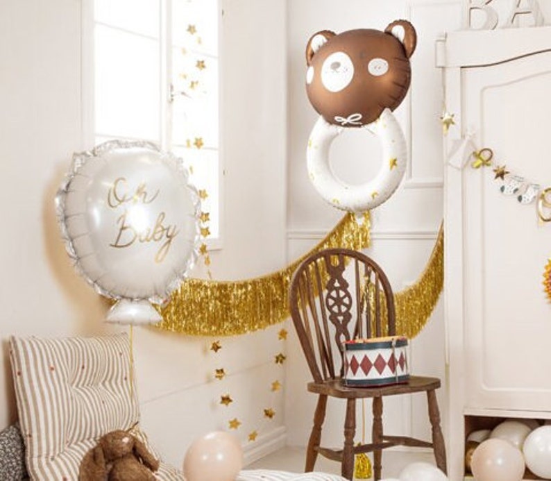 May include: A white balloon with the text "Oh Baby" in gold lettering, a brown bear balloon with a white ring around it, and gold fringe garland.