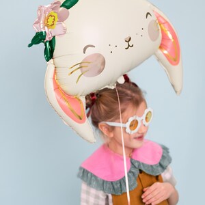 May include: A young girl wearing a brown dress and pink and green collared shirt holds a white bunny balloon with pink ears and a flower on top. The balloon has a cute face with black eyes and a pink nose.
