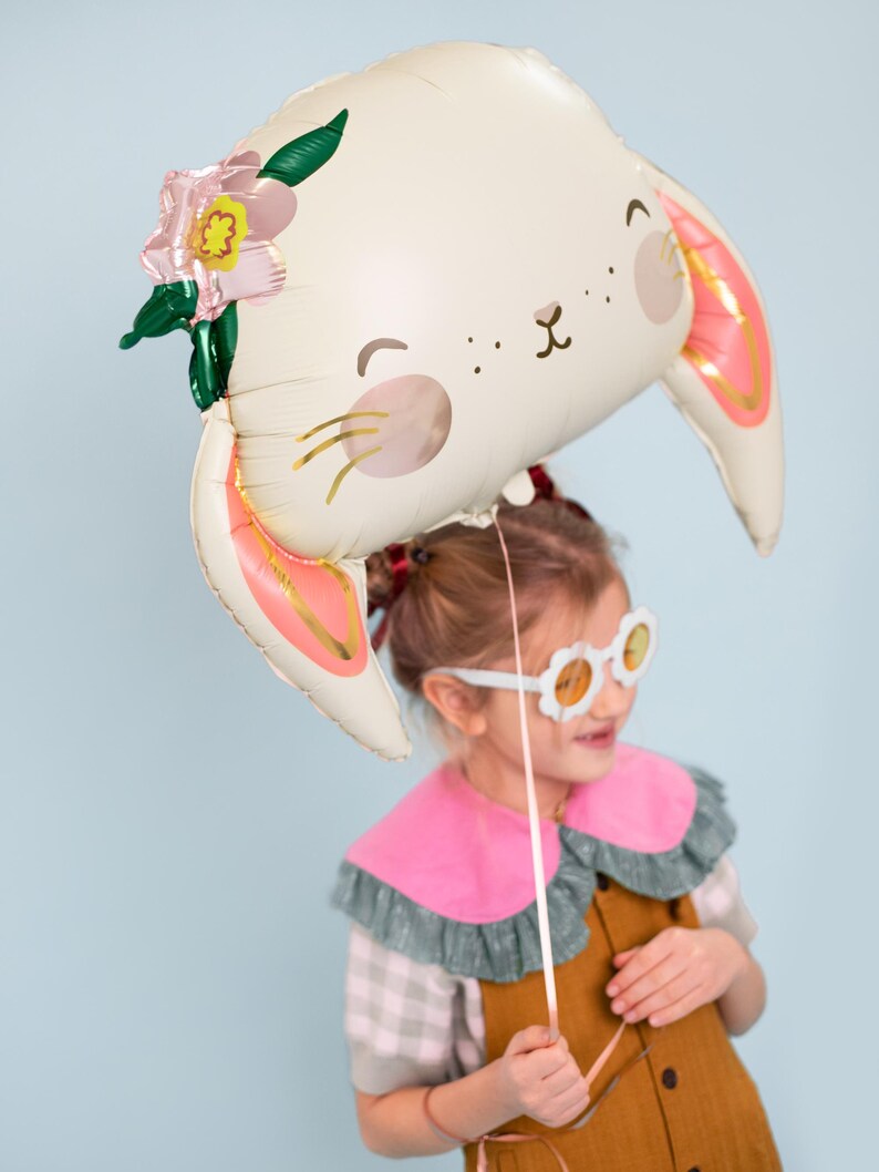 May include: A young girl wearing a brown dress and pink and green collared shirt holds a white bunny balloon with pink ears and a flower on top. The balloon has a cute face with black eyes and a pink nose.