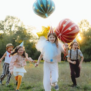 May include: Children running through a grassy field, wearing festive outfits and party hats. Some are holding large, colorful balloons, including a blue and gold star-patterned balloon and a red and white striped balloon. The scene is lit by warm sunlight.