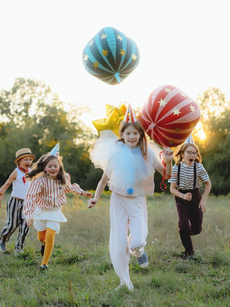 May include: Children running through a grassy field, wearing festive outfits and party hats. Some are holding large, colorful balloons, including a blue and gold star-patterned balloon and a red and white striped balloon. The scene is lit by warm sunlight.