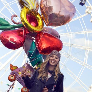 May include: A young woman with long blonde hair is smiling and holding a bunch of colorful balloons in front of a Ferris wheel. The balloons are in the shape of hearts and circles. The woman is wearing a black leather jacket, a bandana, and a black shirt.