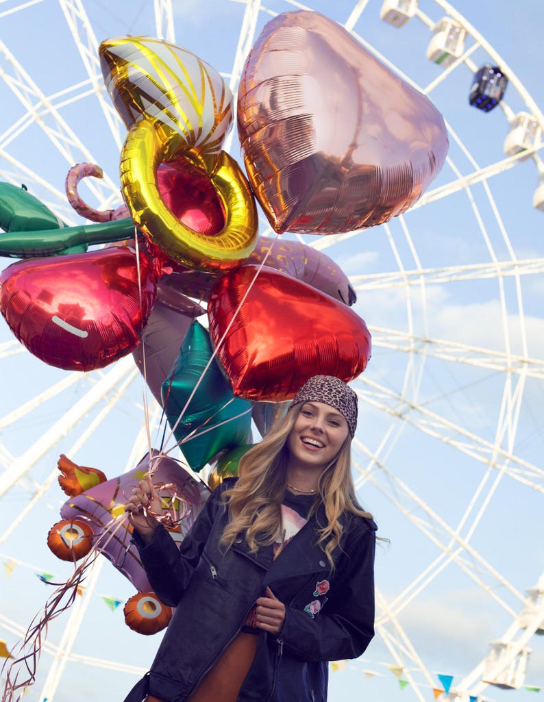 May include: A young woman with long blonde hair is smiling and holding a bunch of colorful balloons in front of a Ferris wheel. The balloons are in the shape of hearts and circles. The woman is wearing a black leather jacket, a bandana, and a black shirt.