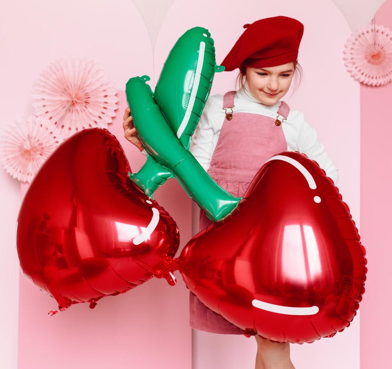 A young girl wearing a red beret, white long-sleeve shirt, and pink pinafore dress is holding a large foil balloon shaped like two connected red cherries with green stems. She is standing in front of a pastel pink background decorated with paper rosettes.