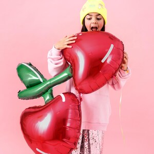 May include: A girl wearing a yellow beanie and a pink sweatshirt holds two large red cherry-shaped balloons with green stems. The balloons are foil and have a shiny finish.