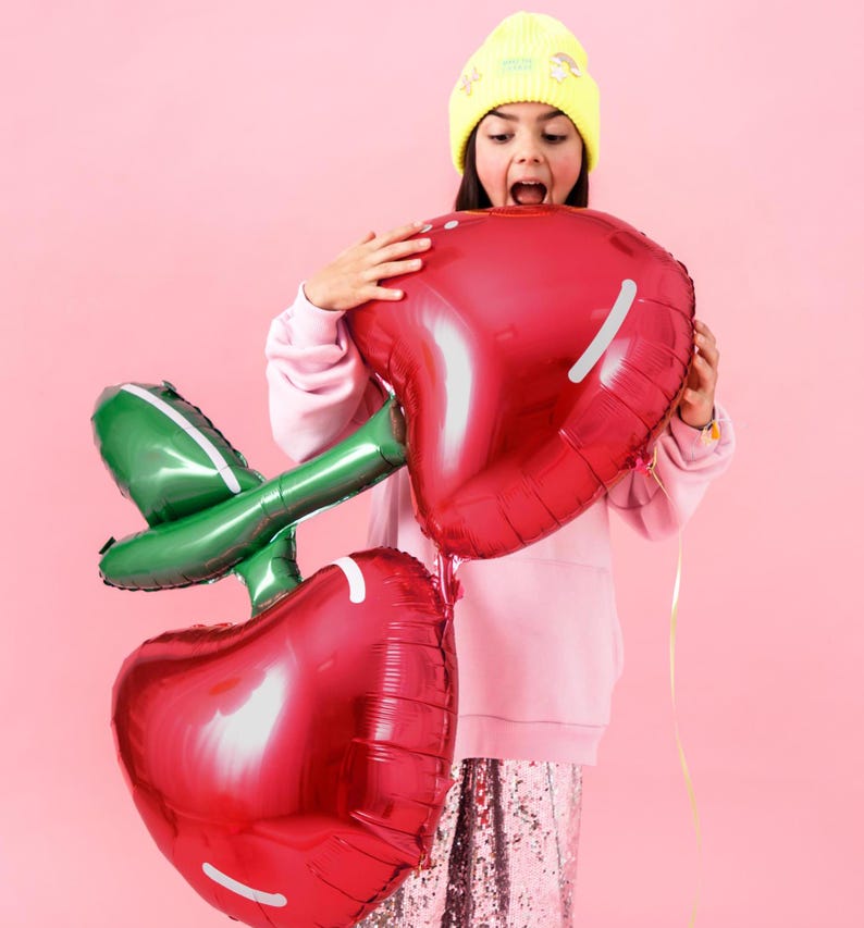 May include: A girl wearing a yellow beanie and a pink sweatshirt holds two large red cherry-shaped balloons with green stems. The balloons are foil and have a shiny finish.