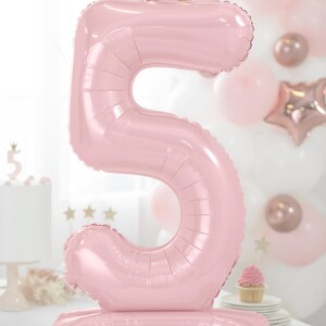 May include: A large pink number 5 balloon with a gold crown on top. The balloon is standing upright on a white surface.