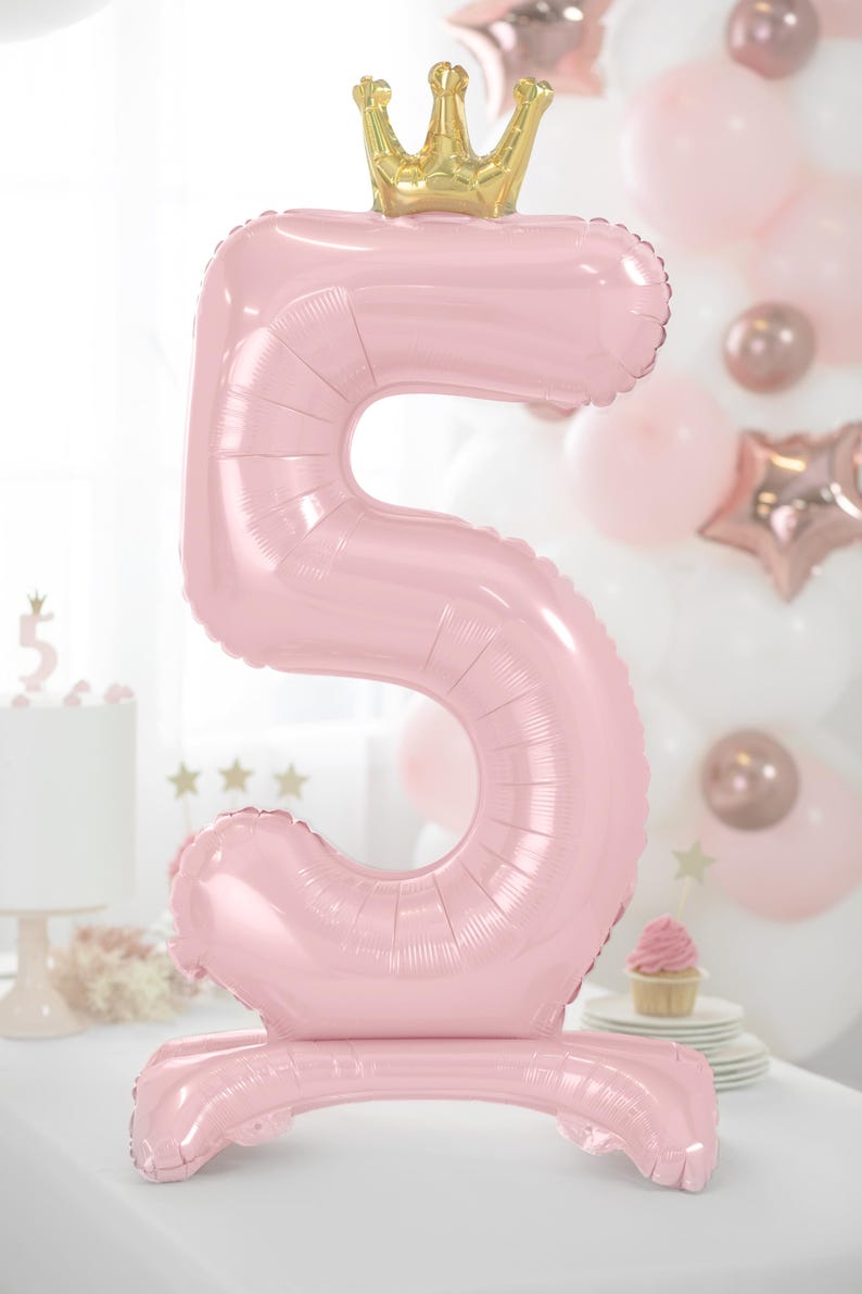 May include: A large pink number 5 balloon with a gold crown on top. The balloon is standing upright on a white surface.