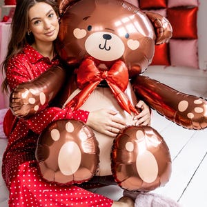 Pu&ograve; includere: Una donna con un pigiama rosso a pois bianchi tiene in braccio un grande palloncino a forma di orsacchiotto di peluche marrone con un fiocco rosso. L'orso ha un viso carino con le guance arrossate.