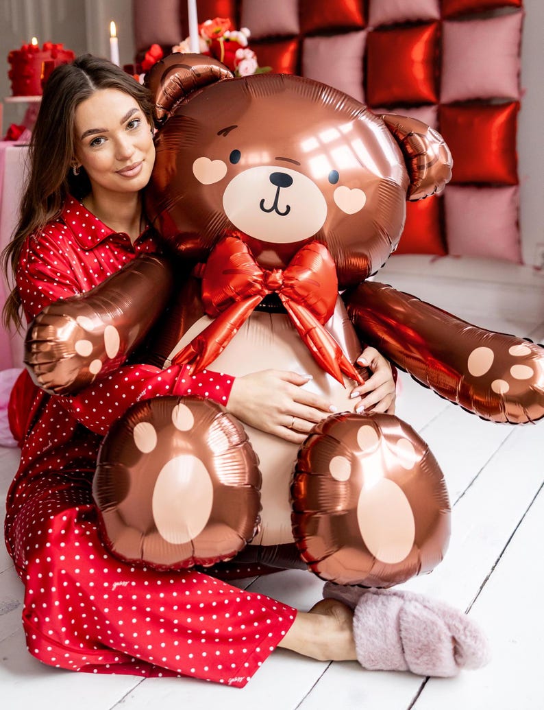Pu&ograve; includere: Una donna con un pigiama rosso a pois bianchi tiene in braccio un grande palloncino a forma di orsacchiotto di peluche marrone con un fiocco rosso. L'orso ha un viso carino con le guance arrossate.