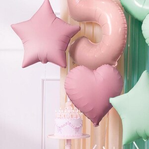 K&ouml;nnte beinhalten: Ein rosa-gr&uuml;nes Geburtstags-Dekorationsset mit einer Torte, Ballons und Kerzen. Die Ballons sind in Form eines Sterns, Herzens und der Zahl f&uuml;nf. Die Torte ist mit wei&szlig;em Zuckerguss und rosa Blumen verziert.