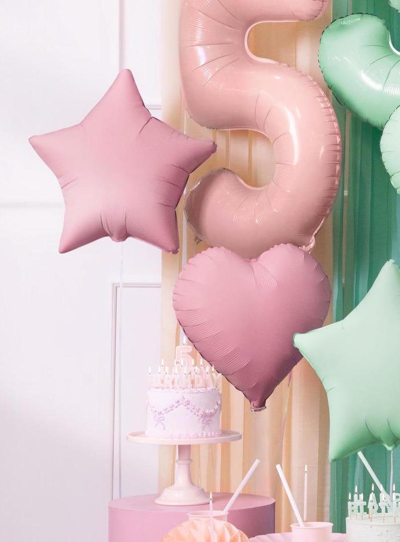 K&ouml;nnte beinhalten: Ein rosa-gr&uuml;nes Geburtstags-Dekorationsset mit einer Torte, Ballons und Kerzen. Die Ballons sind in Form eines Sterns, Herzens und der Zahl f&uuml;nf. Die Torte ist mit wei&szlig;em Zuckerguss und rosa Blumen verziert.