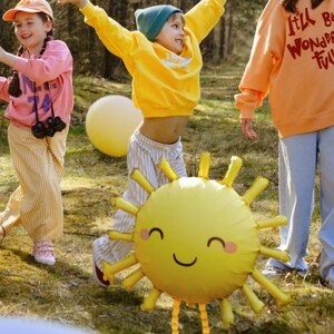 May include: Three children playing outdoors with a large yellow sun balloon. The balloon has a smiling face and two long legs. The children are wearing colourful clothing and are having fun.