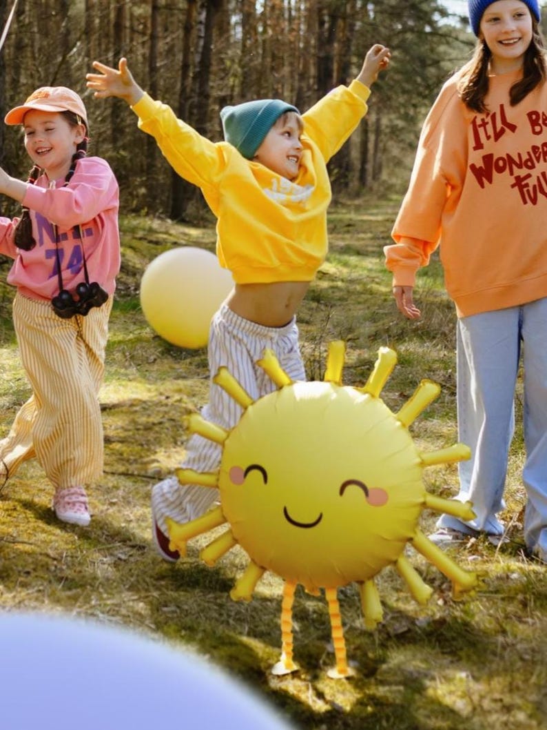 May include: Three children playing outdoors with a large yellow sun balloon. The balloon has a smiling face and two long legs. The children are wearing colourful clothing and are having fun.