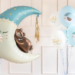 Puede incluir: Un gran globo de luna creciente azul y blanco con un oso marr&oacute;n durmiendo dentro. El globo est&aacute; atado a una cuerda y hay varios globos m&aacute;s peque&ntilde;os azules y blancos con el texto "Baby Boy" en ellos.