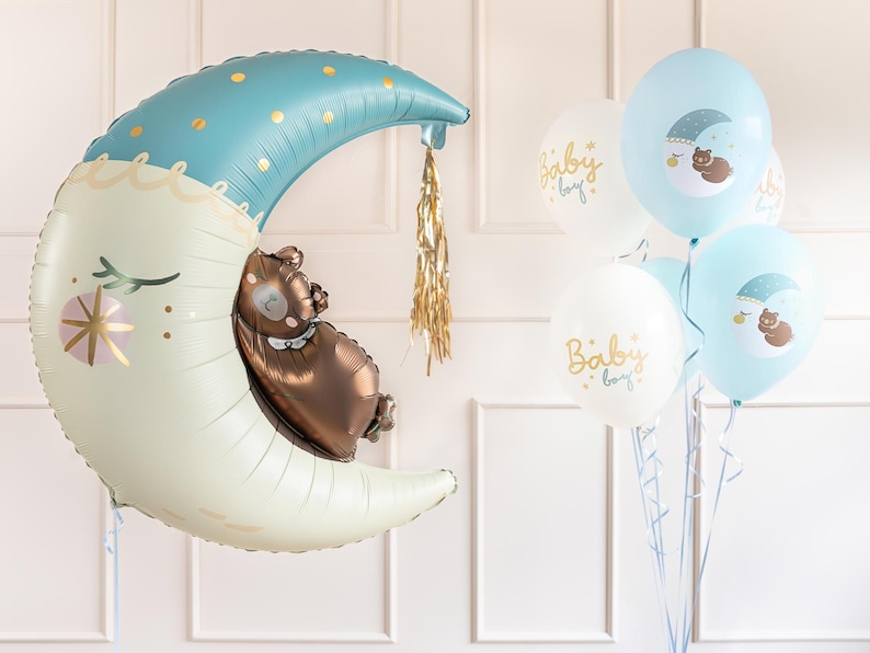 Puede incluir: Un gran globo de luna creciente azul y blanco con un oso marr&oacute;n durmiendo dentro. El globo est&aacute; atado a una cuerda y hay varios globos m&aacute;s peque&ntilde;os azules y blancos con el texto "Baby Boy" en ellos.