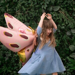 May include: A young girl wearing a blue dress and a pink butterfly balloon. The girl is smiling and holding the balloon above her head. The background is a green wall with lights.