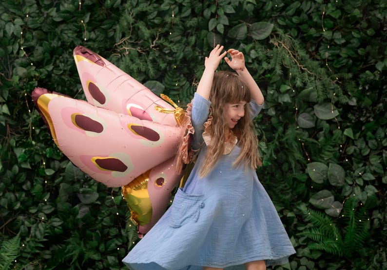 May include: A young girl wearing a blue dress and a pink butterfly balloon. The girl is smiling and holding the balloon above her head. The background is a green wall with lights.