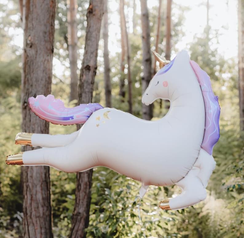 May include: A white unicorn balloon with a purple and pink tail and gold accents. The balloon is floating in a woodland setting.