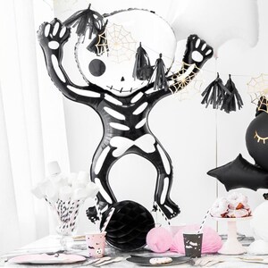 Puede incluir: Un globo de esqueleto negro y blanco con un dise&ntilde;o de telara&ntilde;a. El globo est&aacute; colgado de una guirnalda de borlas negras y doradas. Tambi&eacute;n hay vasos, platos y pajitas de papel negro y blanco sobre una mesa de m&aacute;rmol blanco.