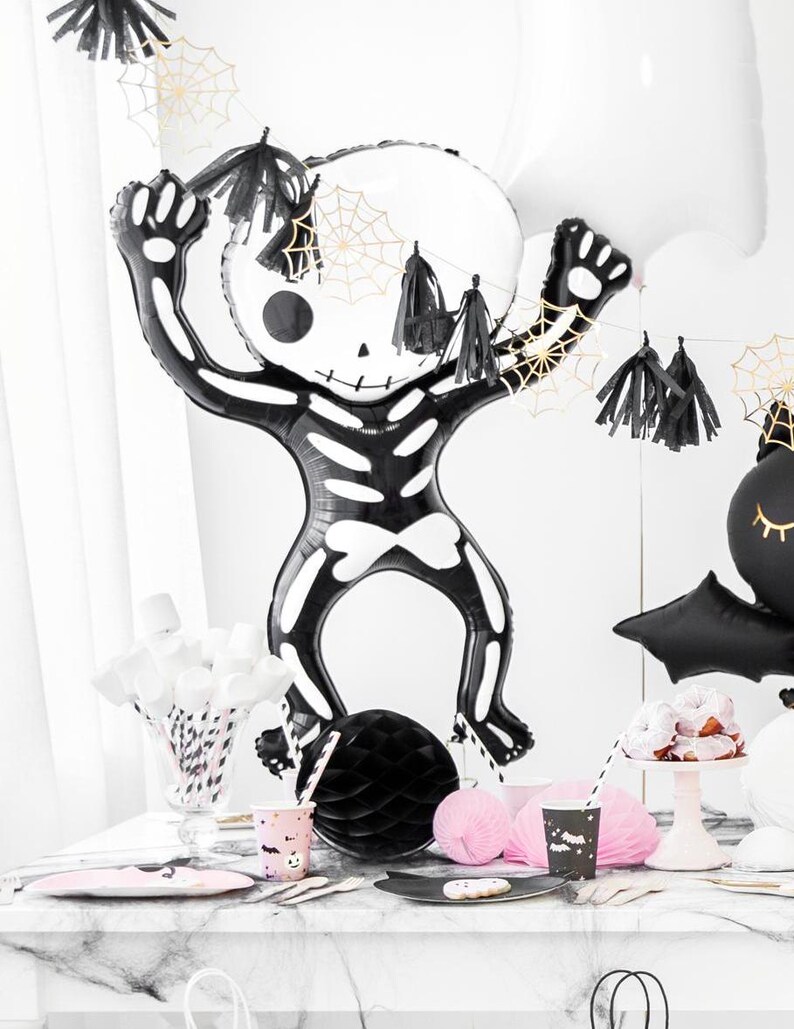 Puede incluir: Un globo de esqueleto negro y blanco con un dise&ntilde;o de telara&ntilde;a. El globo est&aacute; colgado de una guirnalda de borlas negras y doradas. Tambi&eacute;n hay vasos, platos y pajitas de papel negro y blanco sobre una mesa de m&aacute;rmol blanco.
