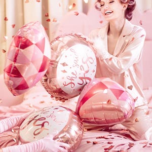 May include: A pink and gold heart-shaped balloon with the text "She said yes" in white. The balloon is being held by a woman in a pink satin robe with her hair in rollers. There are other pink and gold balloons in the background.