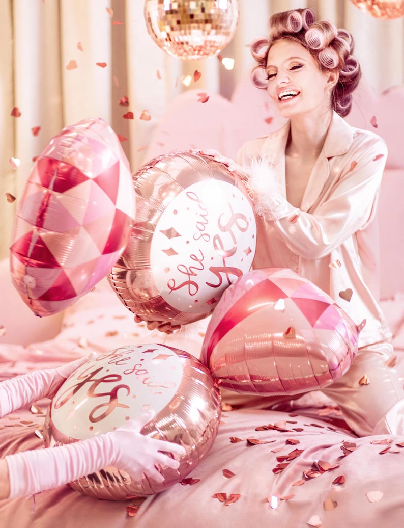 May include: A pink and gold heart-shaped balloon with the text "She said yes" in white. The balloon is being held by a woman in a pink satin robe with her hair in rollers. There are other pink and gold balloons in the background.