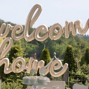 Op de afbeelding: Een grote, beige ballondecoratie met de tekst "welcome home" in cursief schrift. De ballonnen hangen buiten tegen een achtergrond van groene bomen en een heldere lucht. Cupcakes zijn hieronder zichtbaar.