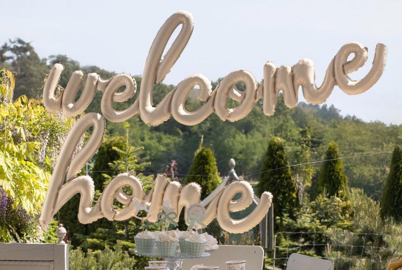 Op de afbeelding: Een grote, beige ballondecoratie met de tekst "welcome home" in cursief schrift. De ballonnen hangen buiten tegen een achtergrond van groene bomen en een heldere lucht. Cupcakes zijn hieronder zichtbaar.