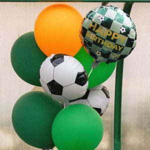 May include: A collection of balloons in green, orange, and soccer ball designs. One balloon reads "Happy Birthday" in gold lettering. The balloons are arranged together, creating a festive and celebratory display.